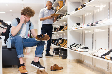 Pensive teenager visiting shoe store in search of new shoes, trying on and choosing stylish loafers while sitting on chair .