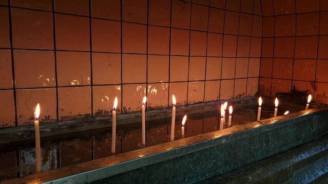 close up footage of numerous candles lid on a concrete platform near altar of a church. Concept image for faith, spirituality, prayer, wish, worship, commemoration events. Candle light blows in wind.