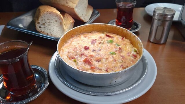 Close up isolated footage of a serving of Turkish menemen, a type of egg omelet made with pepper and tomatoes. It is served in an aluminum pan with sliced white bread and Turkish tea in background.