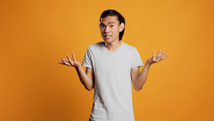 Clueless young person doing i dont know sign in studio, acting uncertain and unsure about answer to question. Asian guy being carefree and casual, acting doubtful over orange backdrop. - obrazy, fototapety, plakaty