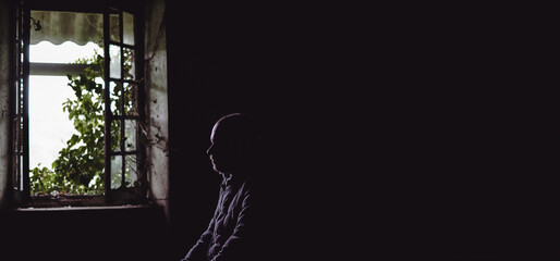 Silhouette of a homeless man in an old uninhabitable house in front of a window. Depression, loneliness, despair, decline, sadness concept. Banner. Place for text.