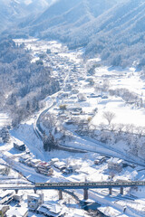 High Angle Landscape of Railway bridge the over the river passing small town in snow day. Pine tree forest mountain with railroad track covered in snow. Beautiful scenic nature in winter season.
