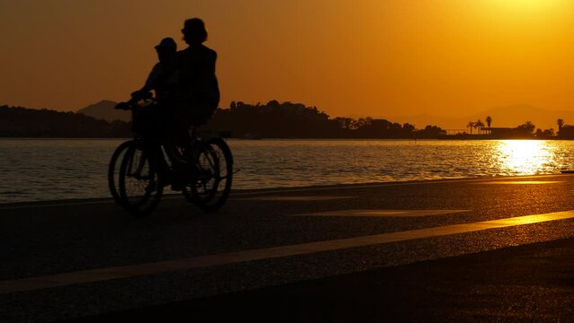 Riding bike silhouette on a walkway. A view of tourist couple ride bikes across the sea shore during evening time in summer.