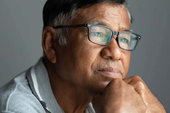 An elderly man wearing glasses with sad expression looks out the window.