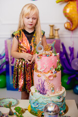 Indoor shot of a rather joyful little girl with blond hair making a wish before blowing out the candle. Celebrates her 6th birthday in a trendy dress, with a huge nautical-themed cake. Happy childhood