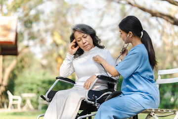 Young asian care helper with asia elderly woman on wheelchair relax together park outdoors to help and encourage and rest your mind with green nature. Use a stethoscope to listen to your heartbeat.