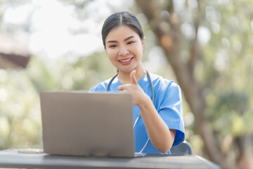 Asian female nurse working outdoor video call with patient online on laptop at desk in park.