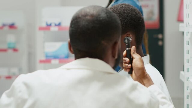 Pharmacy worker consulting man with otoscope tool, otologist doing consultation and ear examination in drugstore center. Young doctor doing otology checkup with male client. Handheld shot.