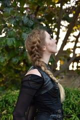 Close up portrait of beautiful female model with blonde plait, wearing black leather catsuit, fantasy assassin warrior.  Posing in forest  tree background