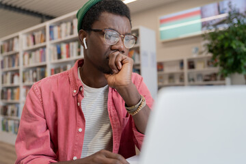 Focused black student guy wearing glasses and wireless earbuds watching online course on laptop, young man freelancer working remotely using free internet in public library. Self-study concept