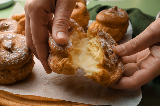 Woman breaking delicious profiterole filled with cream above wooden board, closeup