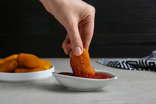 Woman dipping delicious chicken nugget into ketchup at light grey table, closeup
