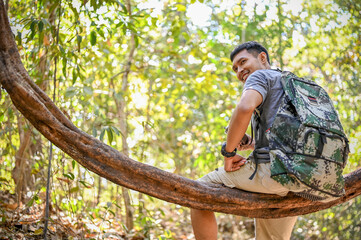 Happy Asian male trekker or traveler sits on a wooden vine, resting while trekking in the forest.
