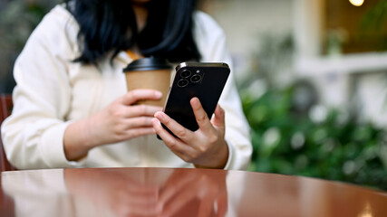 A woman relaxes sitting at the cafe outdoor space, sipping coffee and using her smartphone.