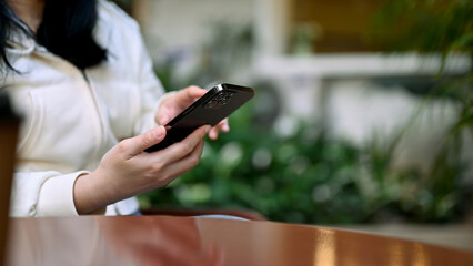 An Asian woman relaxes sitting at the cafe outdoor space and using her smartphone.