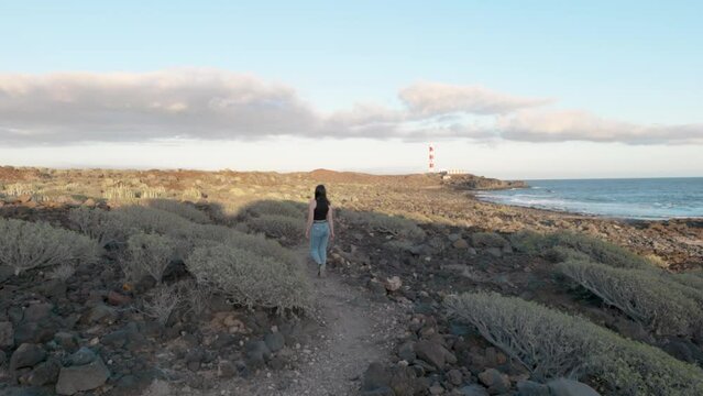 Woman walking through path next to coast and sea, plants, cactuses with lighthouse in background stops and takes a picture, Palm-Ar, Tenerife, Canary Islands, Spain.