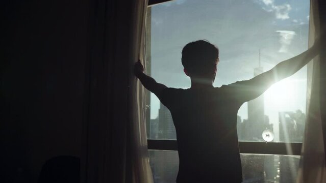 A young man opens the curtains in the morning, raises his arms and stretches. A cinematic man opens large window shades and let the sunlight into the room