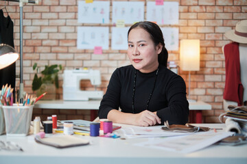 Asian mature female fashion designer is sitting at table, looking at camera and in studio, working with creative and sewing for dress design collection, professional boutique tailor SME entrepreneur.