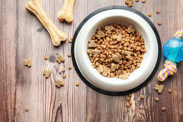 A bowl with dog food, dog treats and toys on a wooden floor.
