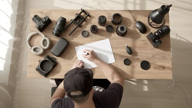 Top down view of man in black cap write sign text THE END on A4 paper. videographer or photographer at his desk writes motivational text in home office with rays of morning sun outside window.