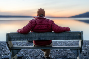 Winter scene of a man with red jacket on a bench looking out over a lake at sunset.  Taking time...