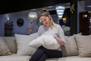 Smiling young woman is sitting on a sofa in a furniture store with a fluffy decorative pillow made of astrakhan in her hands. Interior design and decor. - obrazy, fototapety, plakaty