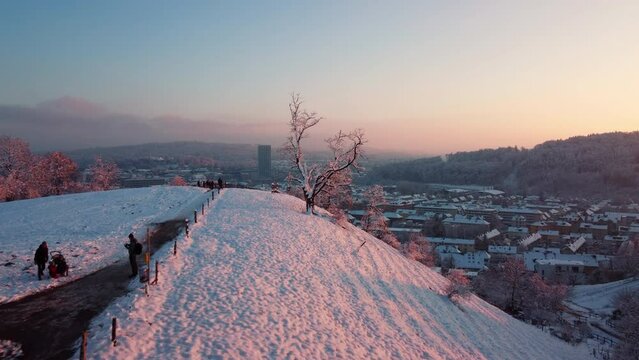 Aerial flight over a snowy hill circling flying towards the skyline of Winterthur, in Switzerland. Setting sun
