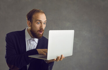 Shocked astonished businessman looking at laptop screen studio headshot against grey wall. Portrait of amazed man employee getting unexpected news. Wow emotion, surprised expression