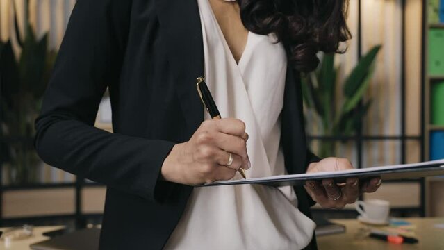 Elegantly dressed business woman signing a document while standing, faceless businesswoman close up parallax