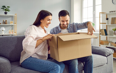 Fototapeta na wymiar Smiling happy young family is sitting together on sofa at home in cozy living room and unpacking large cardboard box. Harmony in relationships, sincere feelings, joy, positivity, satisfaction, fun.