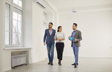 Handsome, bearded male realtor in suit with documents in his hands advises young, happy couple before buying new apartment. Three people are standing in modern spacious living room.