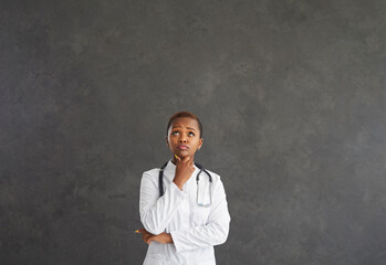Portrait of a serious dark-skinned female doctor with a pensive expression on a gray background. Pensive woman in a medical gown looks at the free space for the text above her head. Banner.