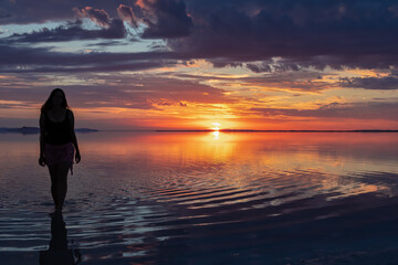 Fototapeta premium Silhouette of woman walking into the sunrise of lake Bonneville Salt Flats, Wendover, Western Utah, USA, America. Dreamy red colored clouds mirroring on the water surface creating romantic atmosphere