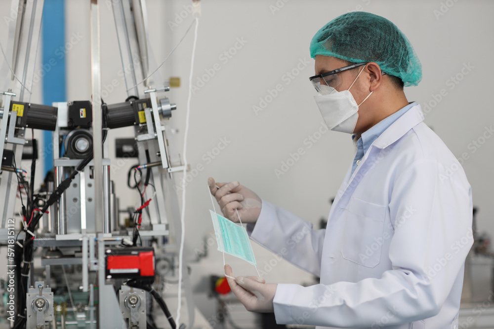 Wall mural Worker man in personal protective equipment or PPE inspecting quality of mask and medical face mask production line in factory, manufacturing industry and factory concept.