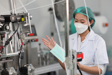 Worker woman in personal protective equipment or PPE inspecting quality of mask and medical face mask production line in factory, manufacturing industry and factory concept.