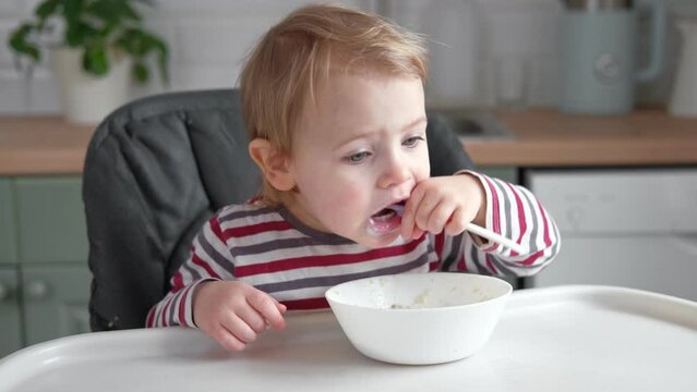 One year old hungry baby girl in striped casual clothes sits at white table in highchair and eats porridge herself with spoon. Blurred dining room background. Healthy eating for kids. Child nutrition
