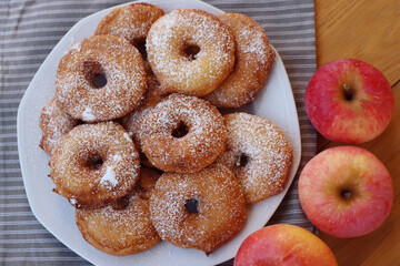Homemade deep fried apple rings with batter sprinkled with powdered sugar ona white plate on wooden table with fresh apples