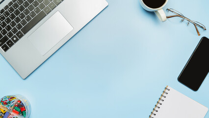 An office desk with a laptop, supplies and a coffee cup on blue background. Top view with copy space