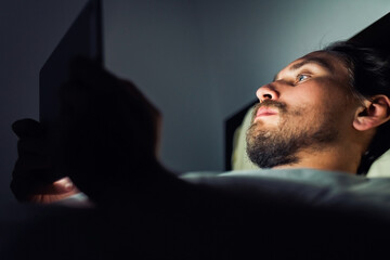 Young handsome and tired man with a beard looking at a tablet at night