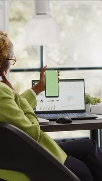 Vertical video: African american woman using greenscreen on phone, working with isolated chroma key display in startup office. Company employee checking blank mockup template on smartphone screen.