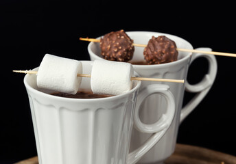 Two beautiful white porcelain cups of hot Chocolate drink with marshmallows and chocolates skewers skewers over wooden table on black background. Winter time. Holiday concept.