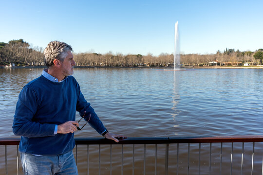 Side view of middle aged bearded man in blue casual sweater leaning on railing while standing against lake with fountain in park