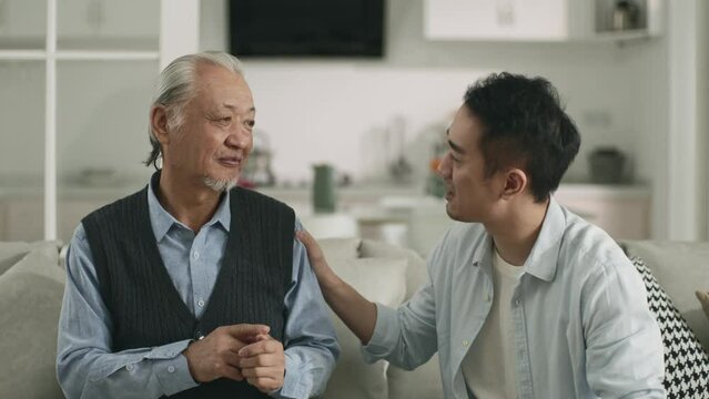 asian adult son sitting on couch at home conversing with senior father happy and smiling