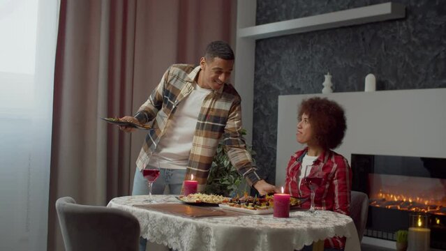 Cheerful caring Attractive black man serving cooked main course to grateful pretty African American woman during romantic dinner, while joyful couple celebrating Valentine's day in domestic room.
