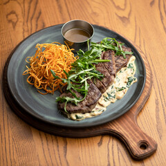 Fried meat with sauce and vegetable side dish on plate. Plate with dish on wooden stand. Meat with creamy sauce, arugula and carrots. Dinner at restaurant. Wooden background. Top view. 