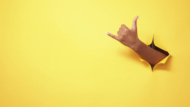 Shaka sign. Hang loose. Call gesture. Female hand showing friendly signal in yellow breakthrough hole ripped paper wall copy space background.