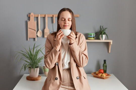 Horizontal shot of joyful young adult woman wearing beige suit standing near table on kitchen at home, holding cup of coffee, keeps eyes closed, enjoying tasty smell of coffee.