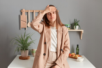 Portrait of sad sick ill young adult woman wearing beige suit standing near table on kitchen at home, keeps hand on forehead, suffering headache, having high temperature.