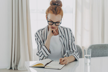 Fototapeta premium Redhead woman in striped shirt takes notes writes down information in notebook