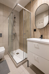 Bathroom with large shower area in corner. Rectangular washbasin on vanity table with white drawers and mirror on wall in black metal frame. Walls made of granite tiles of different textures.